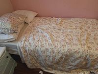 Photo of single bed with floral-patterned comforter, white fitted sheet, and multiple pillows with white pillowcases against pink wall on wooden floor.