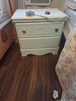 Small bedside dresser shown fully front facing. Painted light green with two drawers and carved floral and geometric designs on front. Metal round knobs on drawers. The dresser is situated between a bed and a chair.