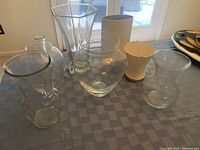 Eight assorted clear glass and ceramic vases on a table with visible details of different shapes and sizes.