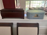 Photo showing both the cherry wood silverware box and the blue decorative metal tin box on a ledge with chairs below and a window in background.