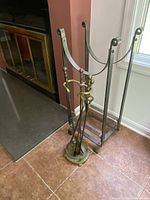 Brass fireplace tool set with holder, poker, tongs, and small shovel next to wrought iron firewood holder by fireplace.
