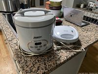 Aroma rice cooker with white and gray plastic body and digital control panel shown on granite countertop.