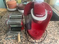 Red Imperia pasta roller with clamp and wooden handle next to red Keurig coffee maker on kitchen counter.