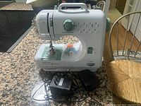 Front view of the Singer Quikfix sewing machine on kitchen counter, showing stitch selector, needle area, and attached cords.