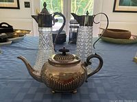Front view showing all three items: two cut glass decanters with silver plated tops and handles, and one silver plated Sheffield tea server teapot, on blue tablecloth.