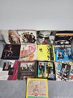 Photo of 13 vinyl 45 RPM records in original sleeves arranged in rows. Sleeve designs visible showing various artists and bands including punk and new wave era covers.
