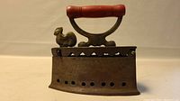 Front side view of vintage metal iron with chicken figurine near handle.