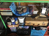 View of lower shelves showing battery charger, kneeling pad with rocks, vintage suitcase, spray cans, black plastic crate with cables and battery charger