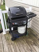 Black propane BBQ grill with closed lid, side burner on right, fold-down shelf on left, propane tank on bottom shelf, blue rope attached.