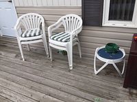 Two white resin deck chairs with green and white striped seat pads on a wooden deck, accompanied by a small white round side table with a blue top and a green planter.