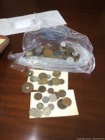 Plastic bag containing a large collection of assorted foreign coins, some loose coins spilled on table and on paper.