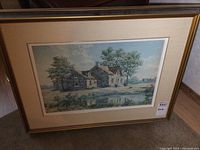 Framed print of a stone house in a landscape with trees, beige matting and gold frame.