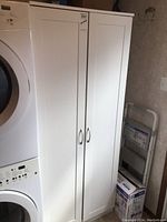 Full front view of white pantry cabinet next to stacked washer and dryer units. Shows two closed doors and silver handles.