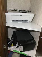 Black HP OfficeJet Pro 8600 printer with cables and power cords nearby on a shelf.