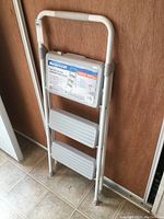 Metal 3-step step stool folded and standing against wall with label visible.