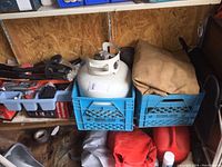 Photo of white propane tank with rust on collar inside blue milk crate, canvas tarp next to it, and other items around on concrete floor.