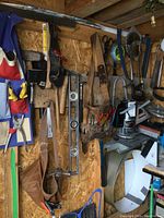 Photo showing various hand tools including hammers, saws, pliers, levels, pry bar, hatchet, tool belts hanging on a pegboard wall.