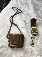 Zenobia camera in leather case with shoulder strap beside Zeiss Ikon light meter on white lace fabric