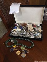Velvet jewelry case opened showing a large assortment of vintage earrings, mostly clip-ons, and pins mixed together. In front, a green jade-like round bead necklace is laid out along with a charm bracelet with multiple charms.