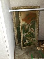 Photo showing both fabric paintings leaning against a wall in a closet space, framed in gold decorative frames.