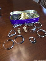 Overview of the vintage ladies jewelry lot with bracelets, bangles, earrings, brooches, compact cases, and watch arranged on a wooden surface with some pieces placed inside a metal purple box.
