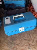 Closed blue Adventure plastic fishing tackle box with white handle and latch on wooden surface.