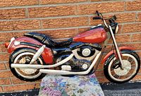 Front and side views of the metal motorcycle wall art against a brick background showing painted red gas tank, black engine, white exhaust pipes.