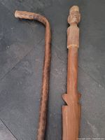 Pair of wood walking sticks shown side by side on dark surface, one with natural curved handle, the other straight with carved details.
