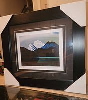Photo showing the framed print with protective corner guards and clear view of the artwork featuring stylized mountains and water.