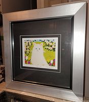 Framed Maud Lewis art print titled "White Cat" showing the full silver frame and detailed image of the white cat with blue eyes and floral backdrop.