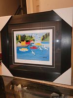 Framed fine art print in dark wood frame with white protective corners, showing bright coastal scene with boats, buildings, seagulls, and trees.