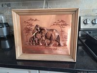 Frontal photo of the vintage framed copper embossed elephant family showing mother and young in a natural setting with trees, placed on a black surface near kitchen appliances.