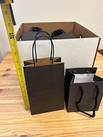 Small black gift bag held next to a measuring ruler showing height approximately 9 inches, placed next to a box of additional bags.
