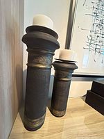 Two black cylindrical candle holders with brass detailing and a candle spike at top, photographed against a wall showing size difference
