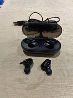 Photo of two black wireless earbuds placed outside an open black charging case with transparent lid and USB charging cable.