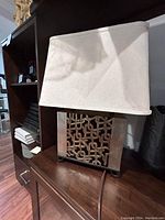 Photo of the lamp on a wooden surface showing its off-white rectangular shade and gold and silver geometric metal base.