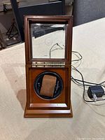 Rotating wooden watch box with glass front panel showing brown watch holder inside, black rotating mechanism, wooden polished case, and power cord adapter attached.