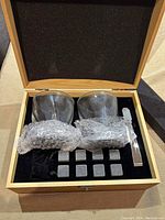 Whiskey glass set contents: two clear whiskey glasses, eight stainless steel ice rocks wrapped in bubble wrap, and a stainless steel tong, presented inside a wooden box with foam padding.