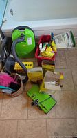 Photo showing various cleaning supplies including a green Kenmore vacuum, red bucket with cleaning bottles, vacuum bags, gloves, duster, handheld device and packaged garbage and lawn bags on tile floor.