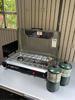 Coleman portable double burner propane camp stove with two propane cylinders on a white table, side and front view