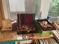 Jewellery display on table showing faux pearl necklaces, black and green bead necklaces, snake head belt, brooches in trays, jewellery box.