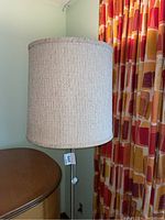 Floor lamp with beige upholstered drum shade and metal tripod base shown near a wooden piece of furniture and colorful curtains.