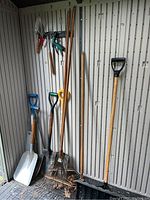 Full view of garden tools including shovels, rakes, and snow shovel standing upright in shed.