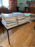Multiple stacks of music books and sheet music on a table in a living room setting, showing a large volume of items.
