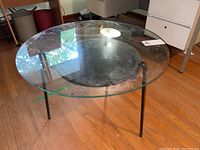 Front-angle view showing clear glass top, black metal legs and visible scratches.