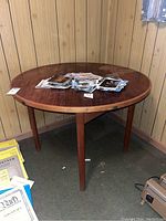 Front view of round wooden table with stained surface, holding a scattered stack of papers, placed on carpeted floor against wood-paneled wall.
