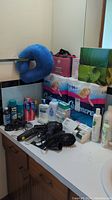 Photo showing unopened packs of Cashmere toilet paper, boxes of No Name facial tissue, various bottled toiletries, hair brushes, hair dryer, and shaver on countertop.