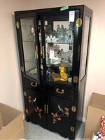 Front and angled view of the tall black Asian style cabinet with glass doors displaying porcelain and decorative figurines inside. Lower section has painted bird and flower designs on the cabinet doors.
