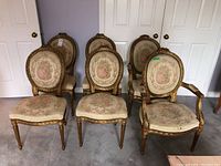 Six antique style wood chairs with upholstered seats and upholstered oval backs with embroidered lady and floral pattern, arranged in two rows against wall, showing wear and fabric damage.