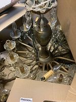 Two vintage chandeliers inside cardboard boxes, showing their metal frames, candle-shaped light bulb sockets, and hanging crystal drops. Some crystals are missing, and the chandeliers need cleaning.
